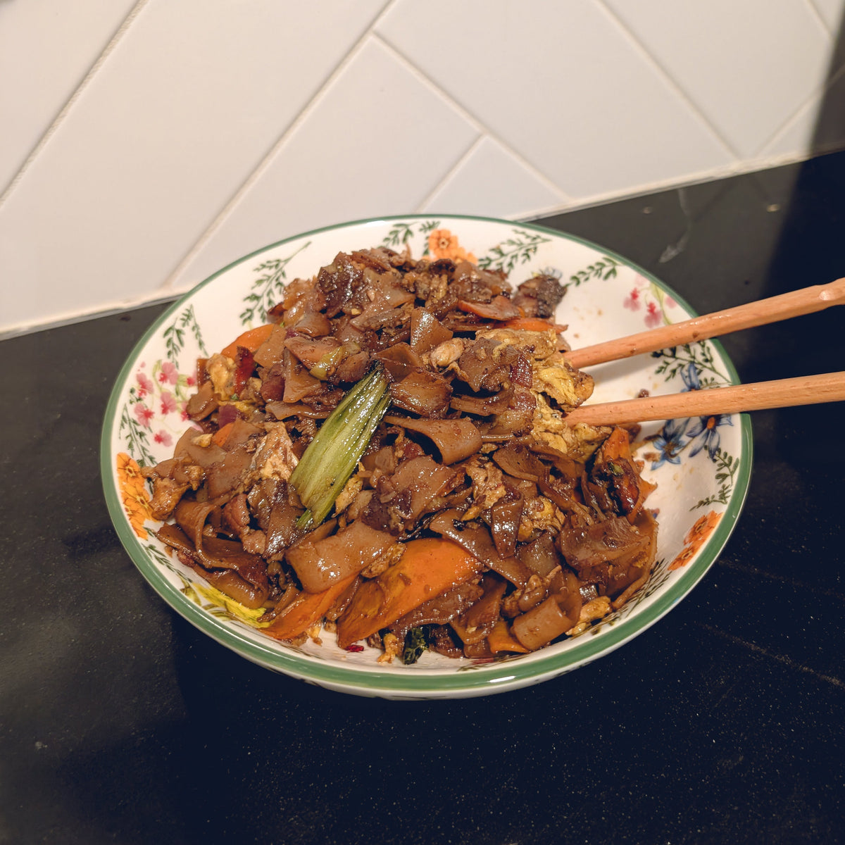 A plate of char-flecked pad see ew noodles with egg and vegetables, finished with Black Garlic Sriracha