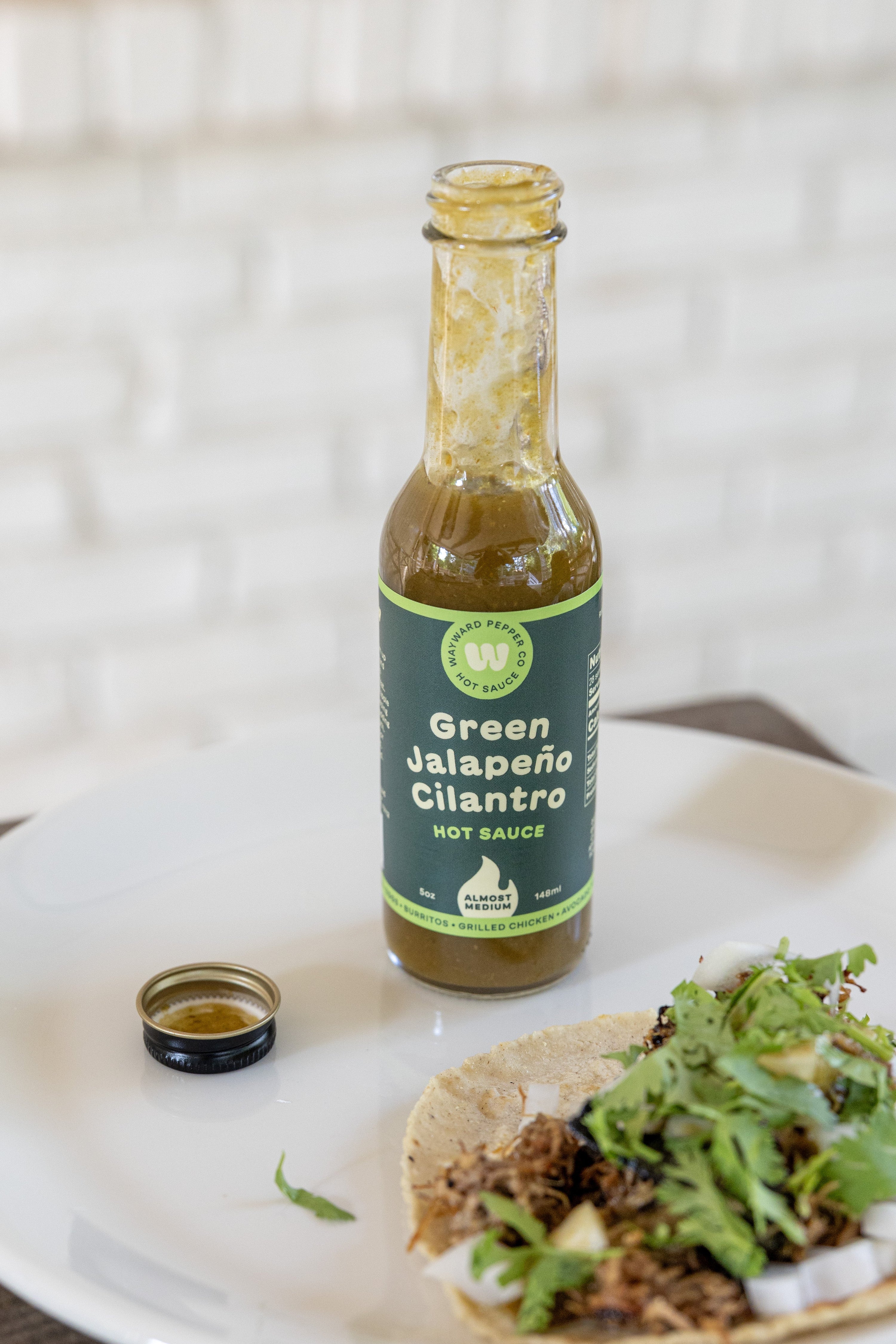 Green Jalapeño Cilantro hot sauce bottle, cap removed, on a table beside a plated dish